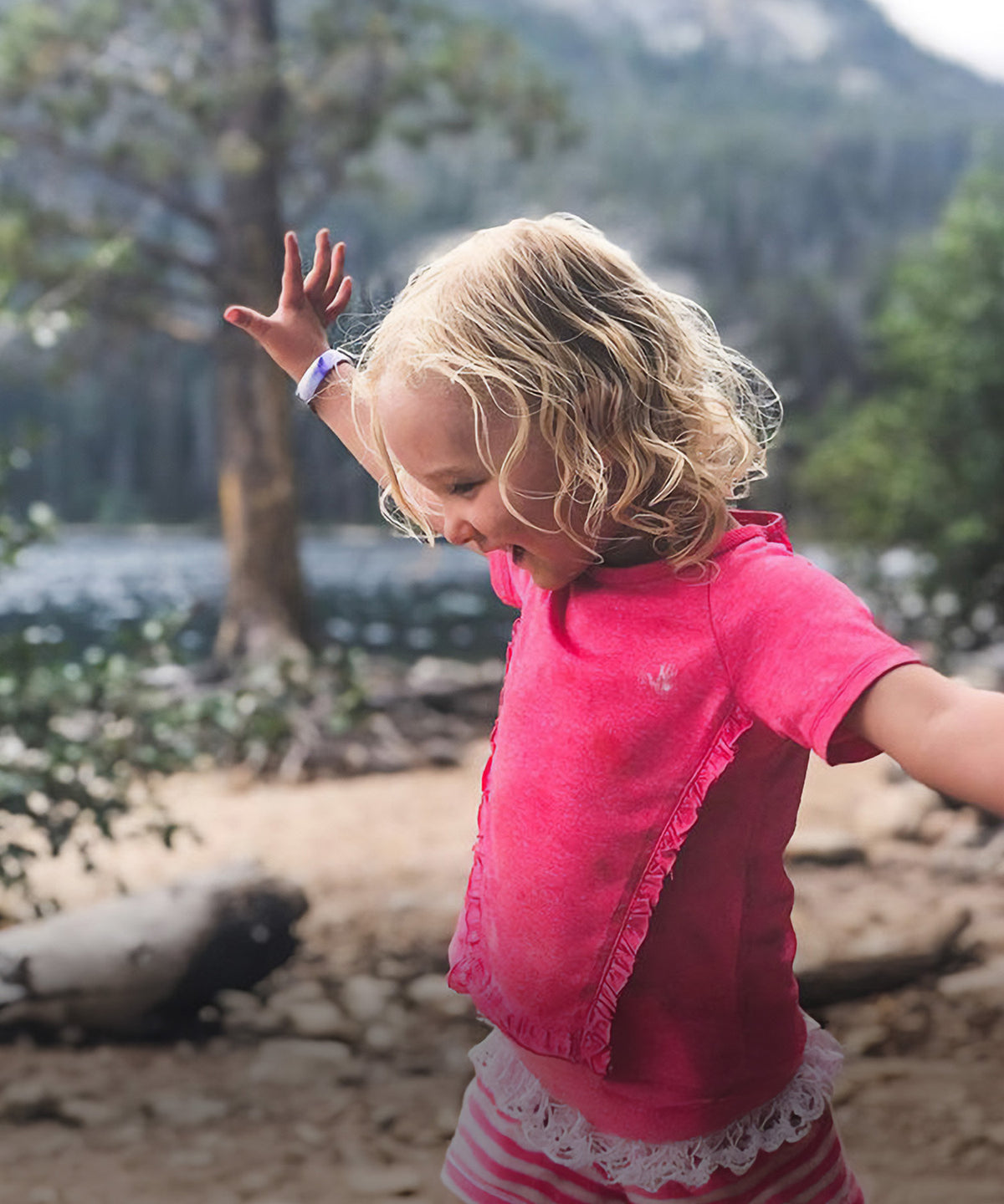 A young girl with blonde curly hair wearing a bright pink short-sleeve top and striped shorts, standing outdoors in a wooded, mountainous environment wearing ROADiD.