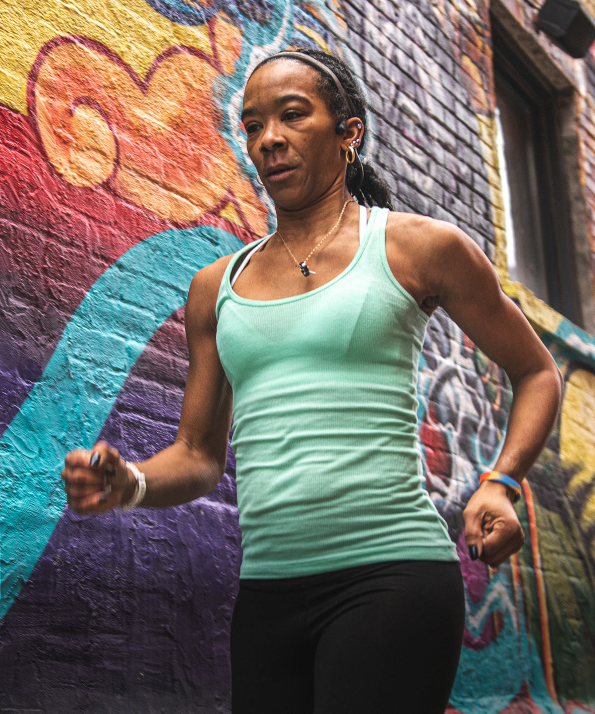 A woman jogging in a mint green athletic tank top and black leggings in front of a colorful graffiti-covered brick wall wearing ROADiD.
