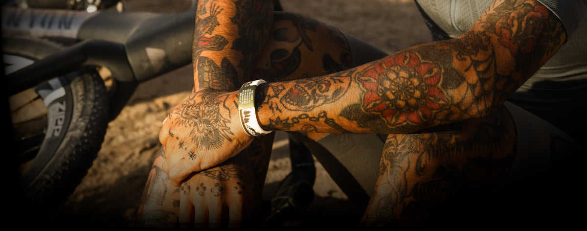 A close-up of a tattooed person's hands and arms wearing a white silicone wristband with a green label and a silver chain bracelet.