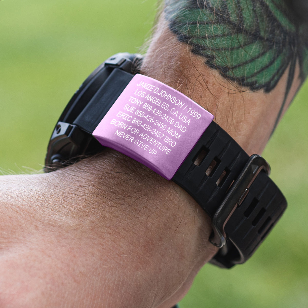 A close-up of a person's wrist wearing a black digital watch with a bright pink rectangular nameplate. The nameplate is printed with white personalized text including 'JAMES D JOHNSON 1980 LOS ANGELES, CA USA' and other memorial details.