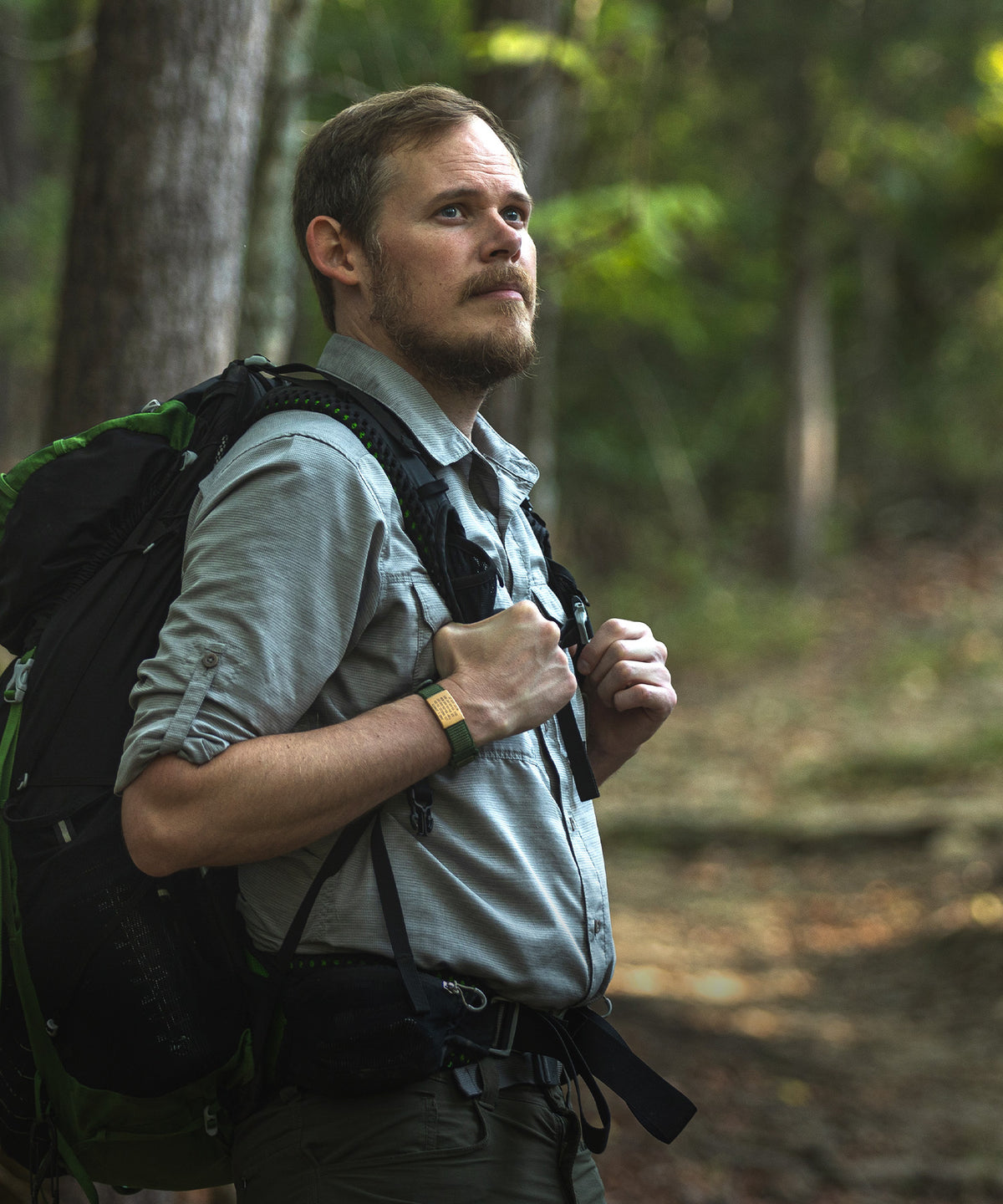 A man hiking in a forest, wearing a light gray button-up shirt, dark hiking pants, and a large black and green backpack wearing ROADiD.