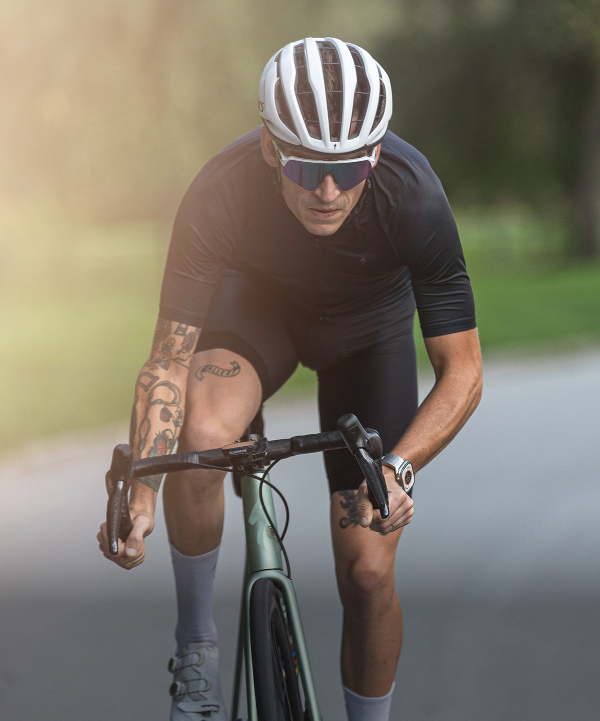 A male cyclist in a black cycling jersey and matching shorts, wearing a white helmet and sunglasses, riding a road bike on a blurred outdoor road wearing ROADiD.