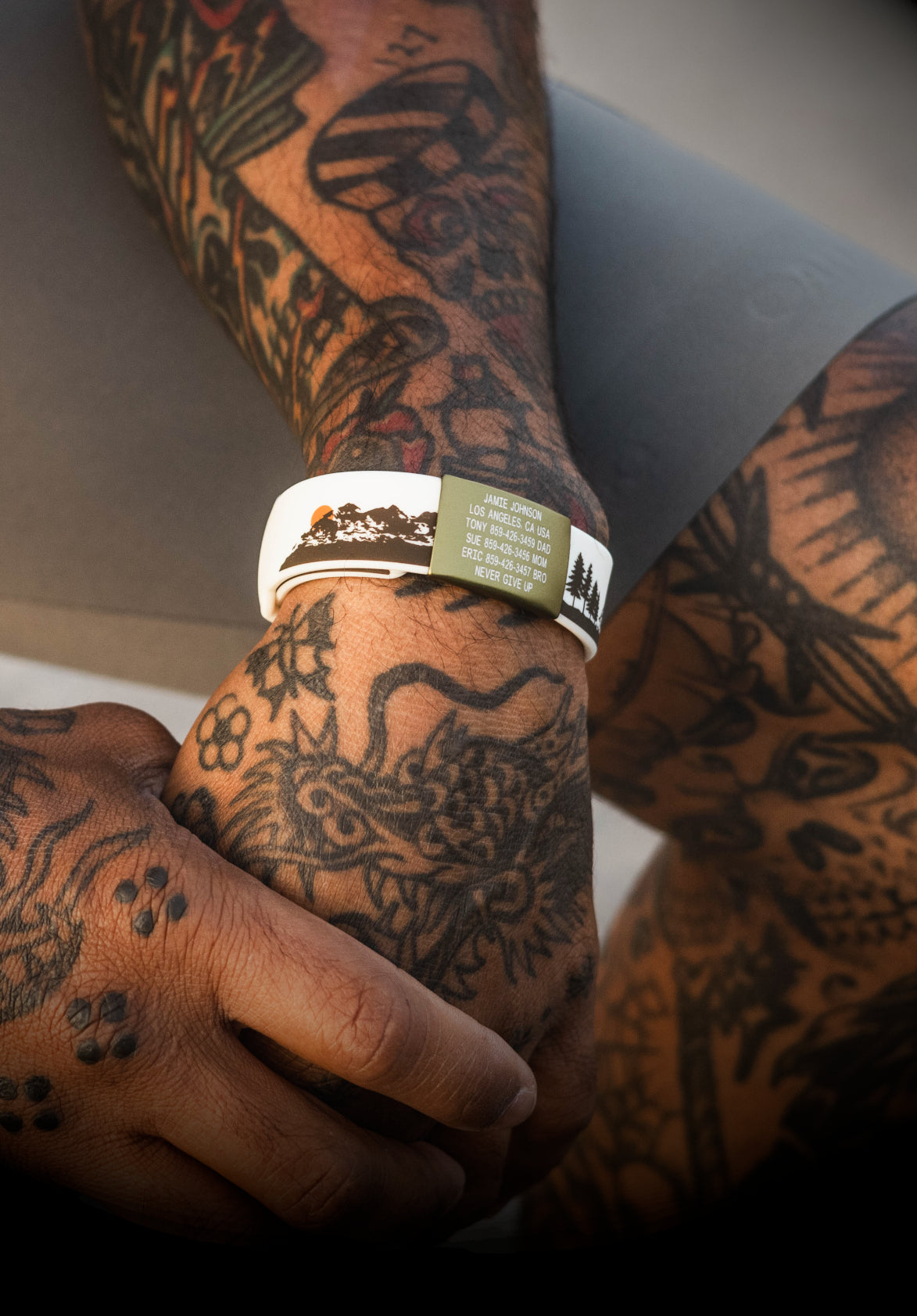 A close-up of a tattooed person's hands and arms wearing a white silicone wristband with a green label and a silver chain bracelet.